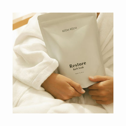 Minimal lifestyle image of Keem Keem Restore Bath Soak pouch held in hands while wearing a white robe, highlighting its calming magnesium formula.