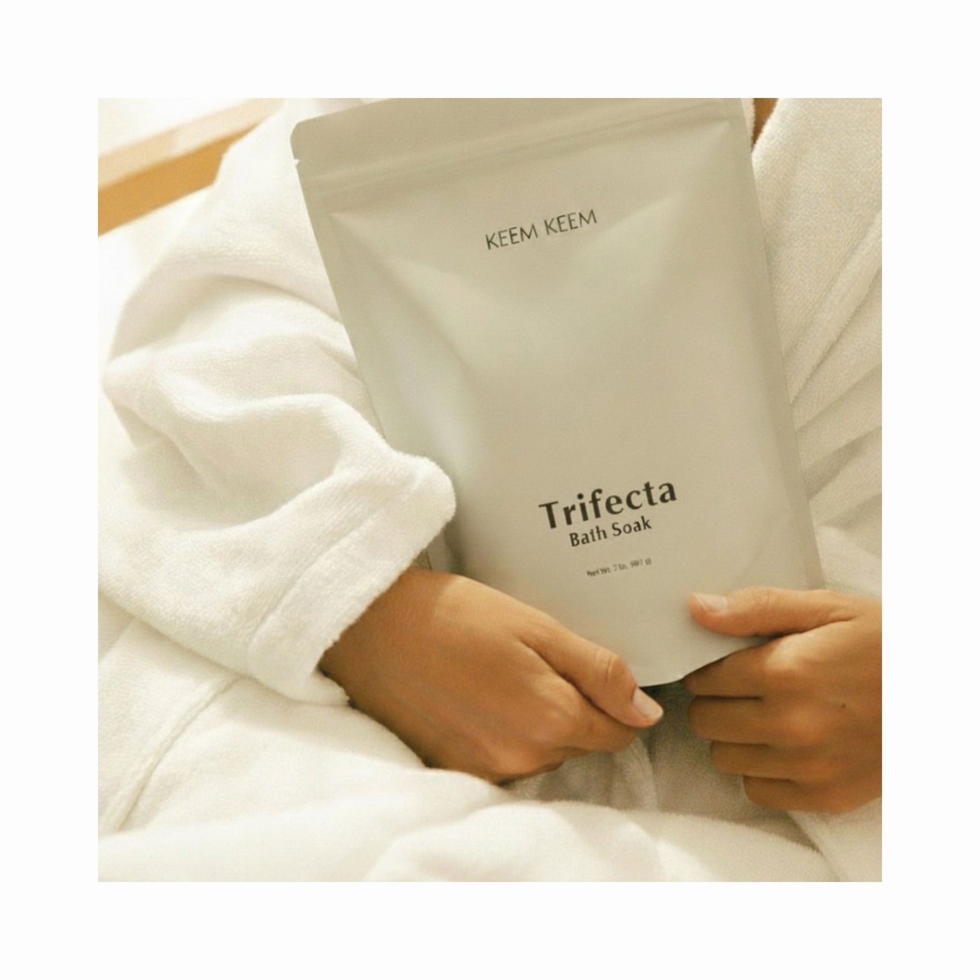 Minimal lifestyle image of Keem Keem Trifecta Bath Soak pouch held in hands while wearing a white robe, showcasing its extra-strength triple-salt recovery blend.