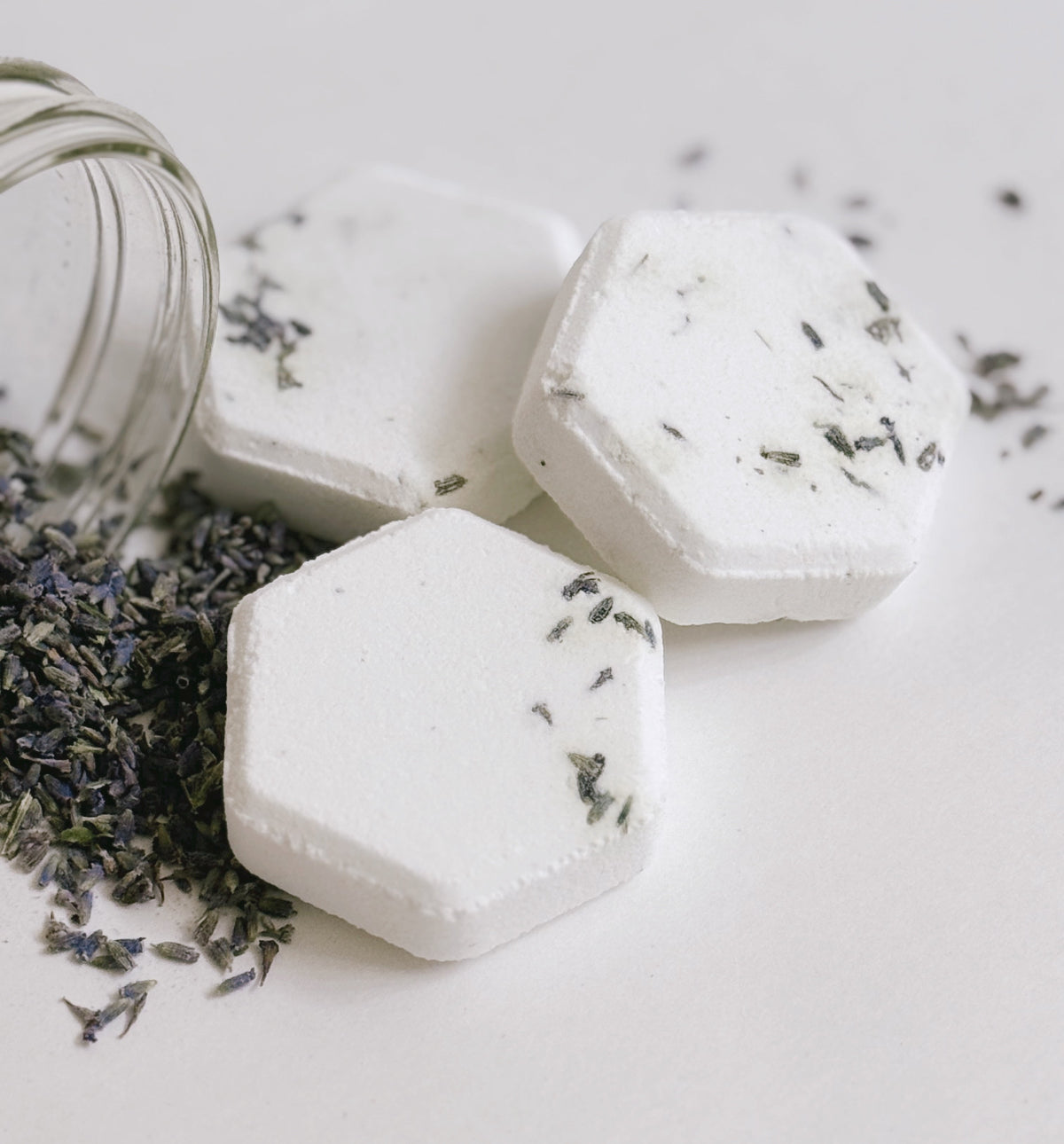 Hexagon-shaped Keem Keem Lavender Shower Steamers with dried lavender buds, shown spilling from a glass jar on a white background
